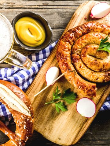 German bratwurst, pretzel, beer, and mustard on a wooden board with a Bavarian blue-white Oktoberfest napkin.