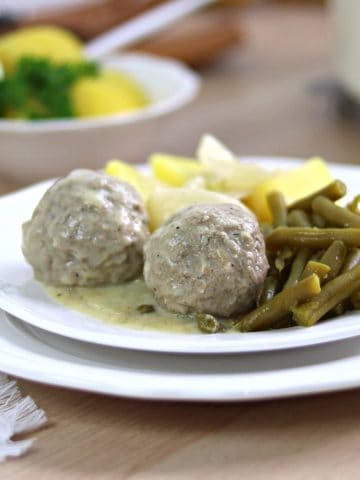 Königsberger Klopse with green beans and boiled potatoes on a white plate. Serving of Salzkartoffeln in the background.
